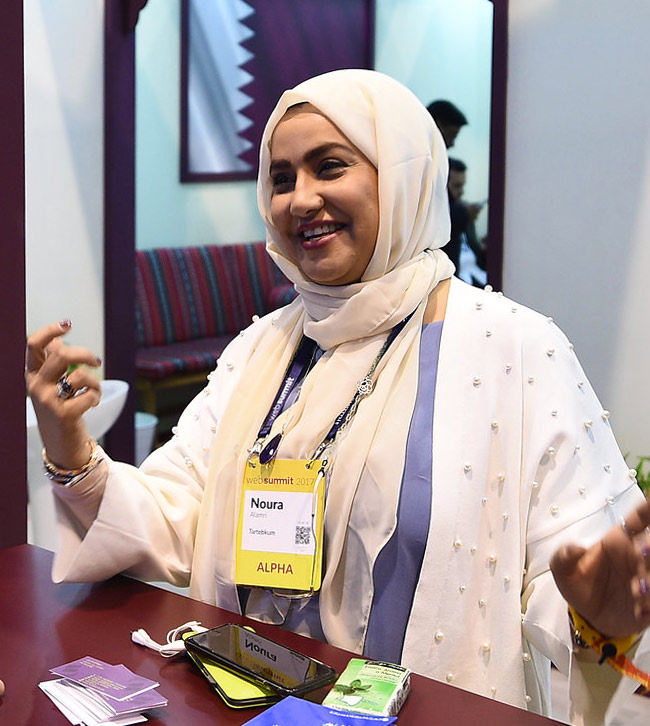 A smiling woman wearing a hijab stands at a wooden counter. She gestures with both hands. Two mobile phones and a stack of business cards are on the counter in front of her. In the background, the Qatari flag hangs on a wall in an adjoining room.
