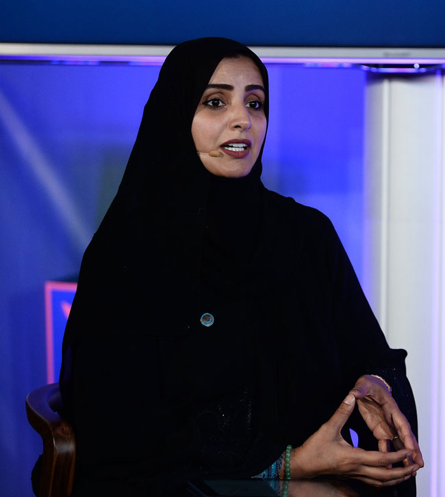 A woman in a hijab sits in a wooden chair at a table. Her fingertips are touching as she makes a tent-like shape with her hands, which are resting on the table. She is wearing a headset mic and appears to be speaking. The woman is Aisha Bin Bishr of Smart Dubai.