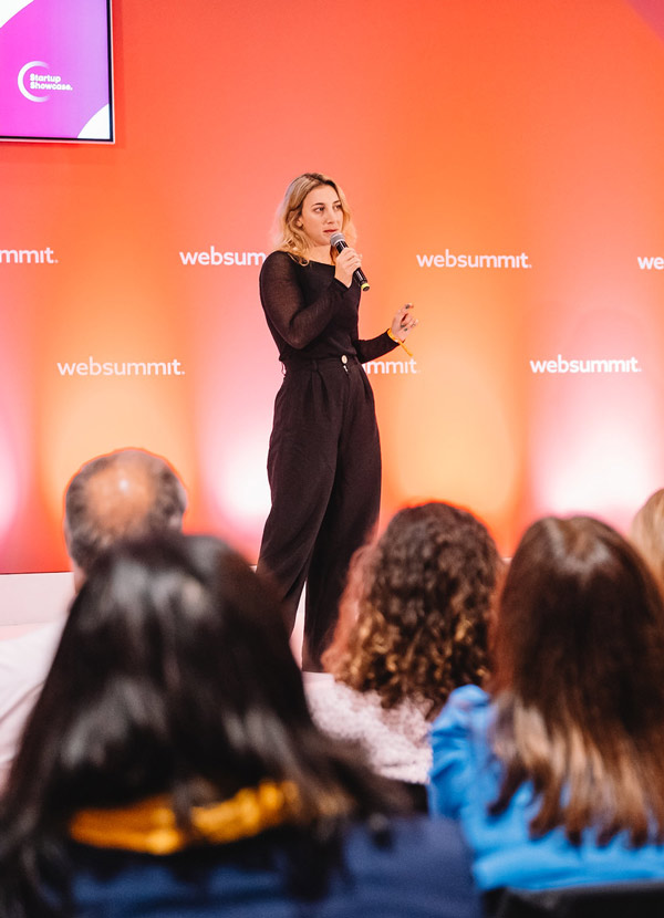 A person stands on a stage. They hold a microphone in their right hand, and their left hand is raised slightly as if emphasising a point. They appear to be speaking. The wall behind them prominently features the Web Summit logo in several places. People in the foreground of the image face towards the stage, and appear to be seated. Only the backs of their heads are visible.