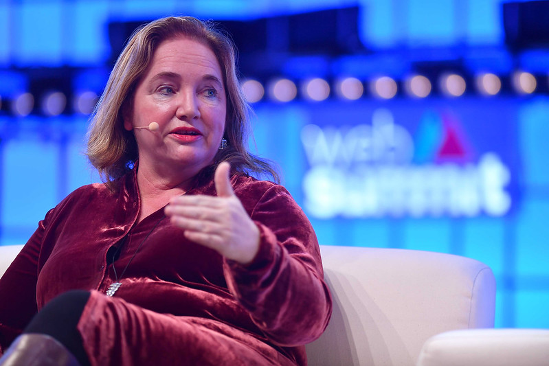 A person (co:collective co-founder and CEO Rosemarie Ryan) sits in an armchair. Their left leg is crossed over their right, and they're gesturing with their left hand. They're wearing a headset mic and appear to be speaking. The Web Summit logo is visible behind them.