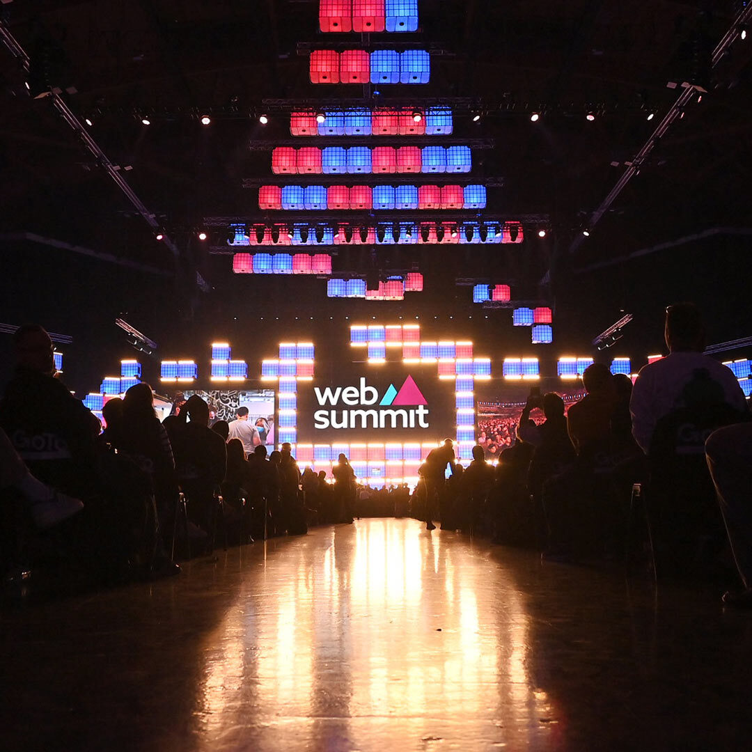 A large stage viewed from far away. The photo is taken from a low angle along an aisle between seats, with the stage's reflection showing on the floor. To either side of the aisle, silhouettes of seated people are visible. In the centre, hanging above the stage, is a large Web Summit logo.