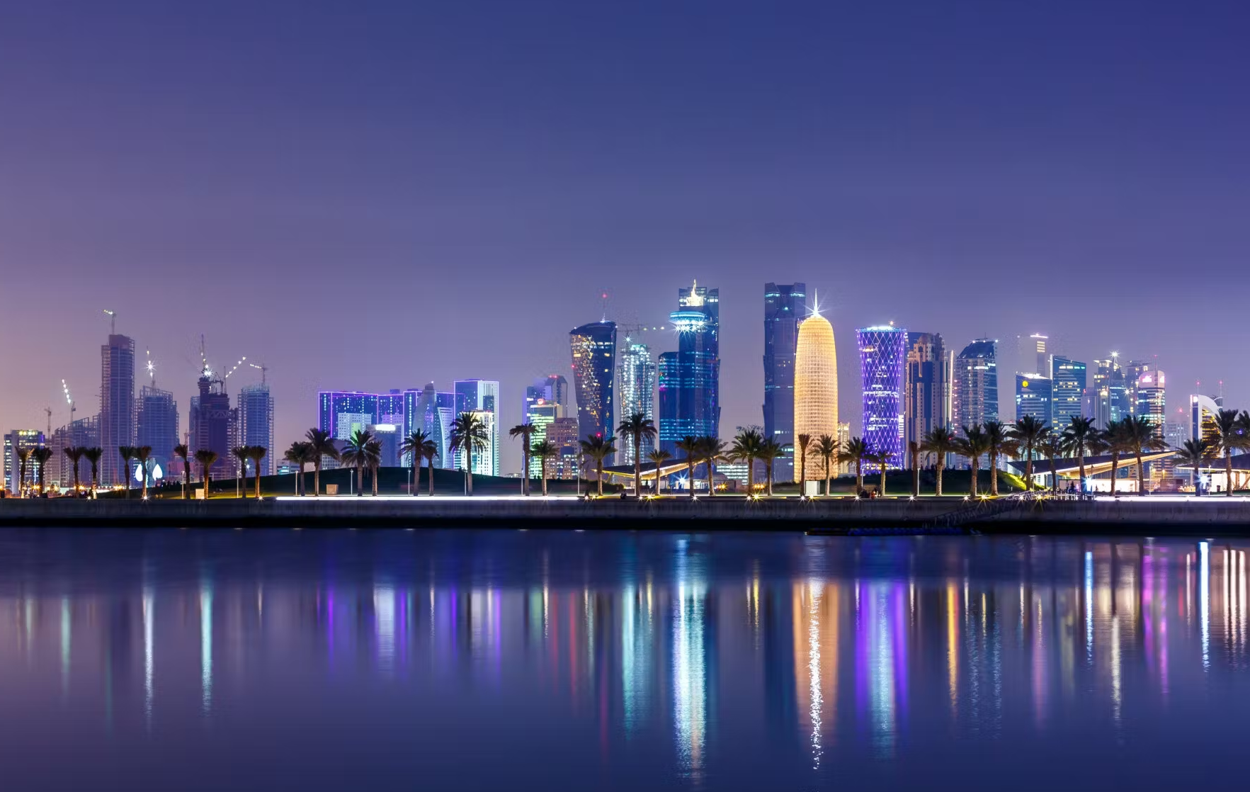 Night time. A brightly lit skyline, viewed across calm waters, featuring several modern glass skyscrapers. There is a row of palm trees where the water meets the shoreline.