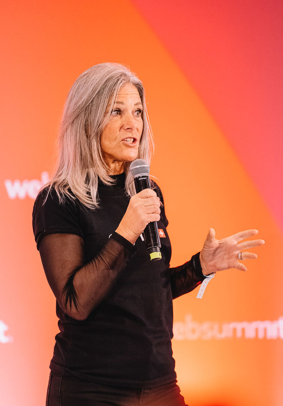 A photograph of a startup pitch on the Startup Showcase stage. The person is holding a microphone with their right hand and using their left hand to gesture. They appear to be speaking to a crowd.
