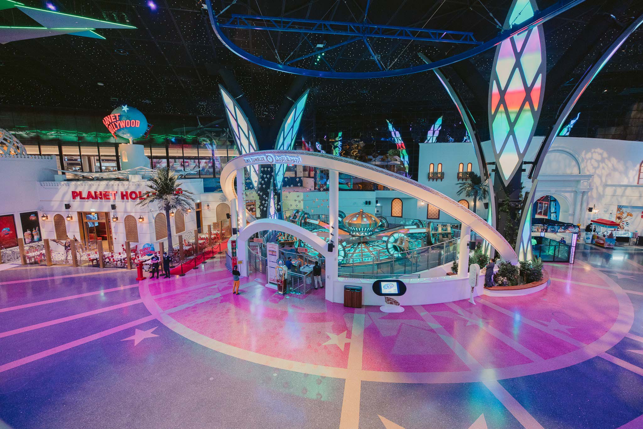 An aerial view of a spacious hall. On the left, a Planet Hollywood restaurant designed in a Middle Eastern architectural style. Next to Planet Hollywood, two people (who appear to be security guards) stand at the end of a red carpet lined with velvet rope. In the centre is an amusement ride called Time Tumbler. It appears to be in the octopus style, with eight arms that revolve around a central axis of rotation while moving up and down in a wavelike motion. To the right of the Time Tumbler is a large door that appears to lead to another room. To the right of this is a concession stand. The ceiling of the space is painted to look like the night sky.