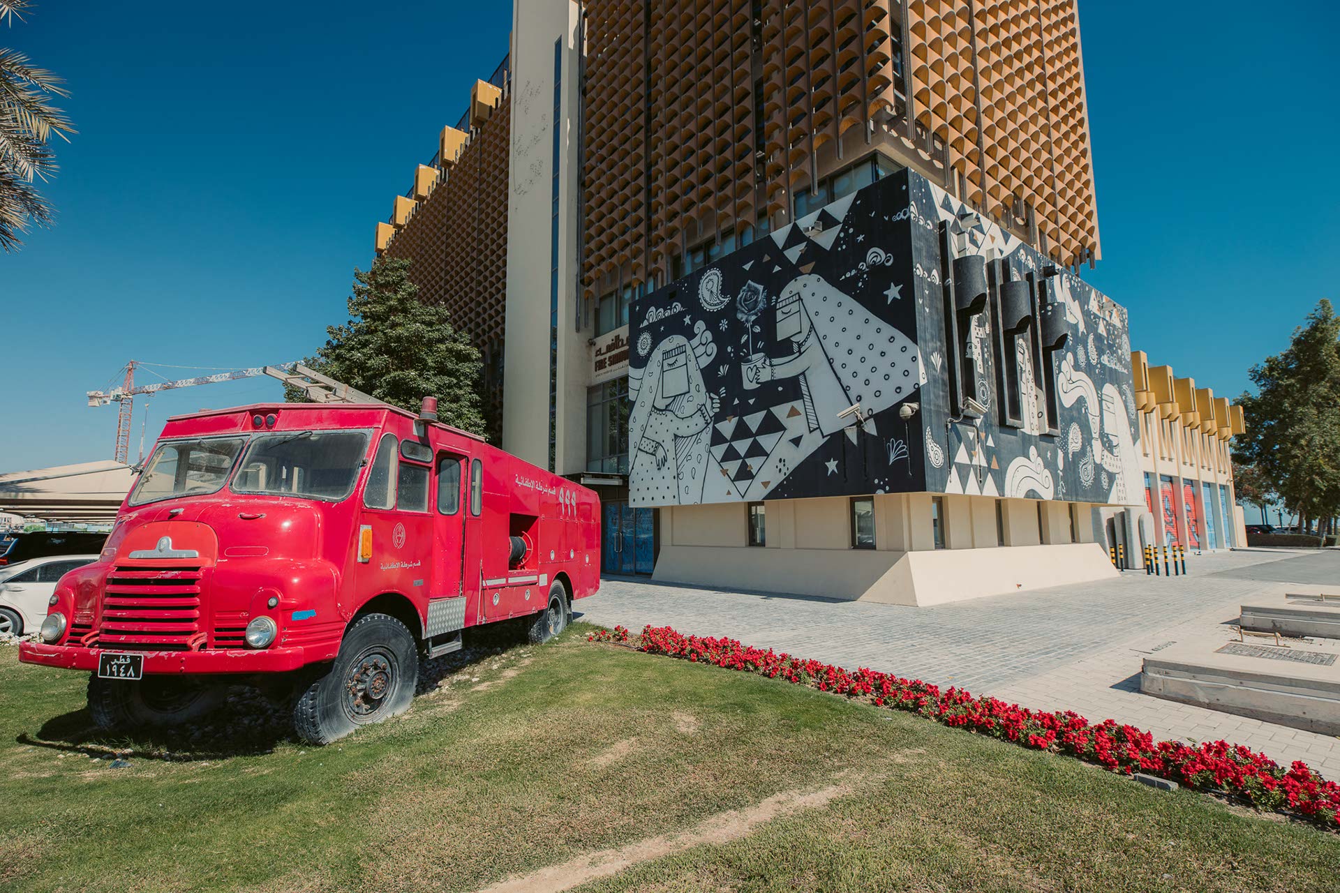 A decommissioned fire truck sits on grass in front of a multi-storey building with a modern facade. This is Fire Station, a contemporary art space. A cartoon-style mural of veiled women with flowers, bells and more wraps around the corner of the building closest to the fire truck. A walkway separates the truck and the building. A construction crane is visible in the background. It is daytime.