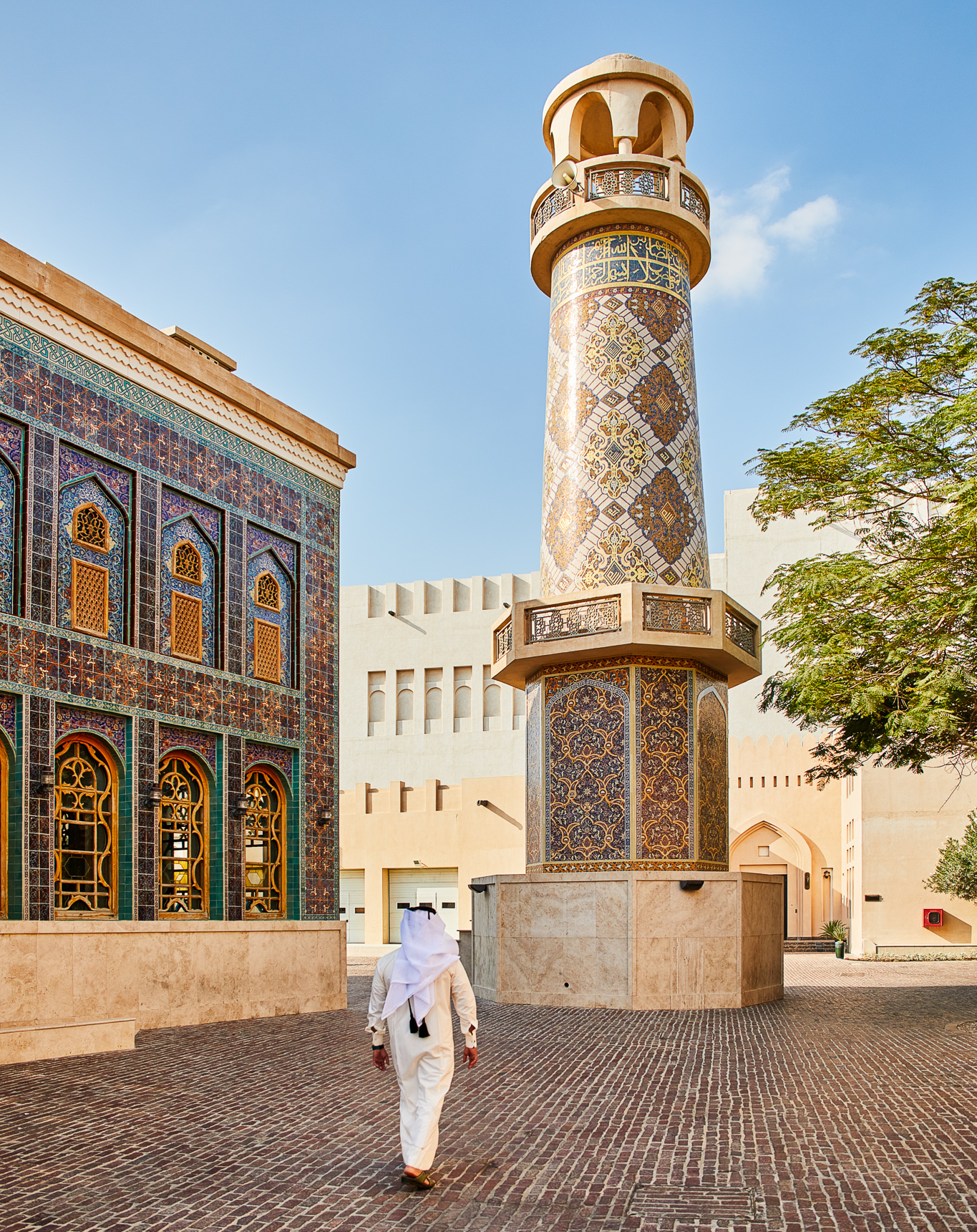 A person walks away from the camera across cobbled stone. On their left is a two-storey building with marble and tile decor on the exterior walls. Decorative screens cover the windows on the upper level. In front of the person is a round minaret with a loudspeaker attached near the top. The minaret has tile and marble decoration. Behind the minaret are buildings that are painted plainly. To the right is a leafy tree.