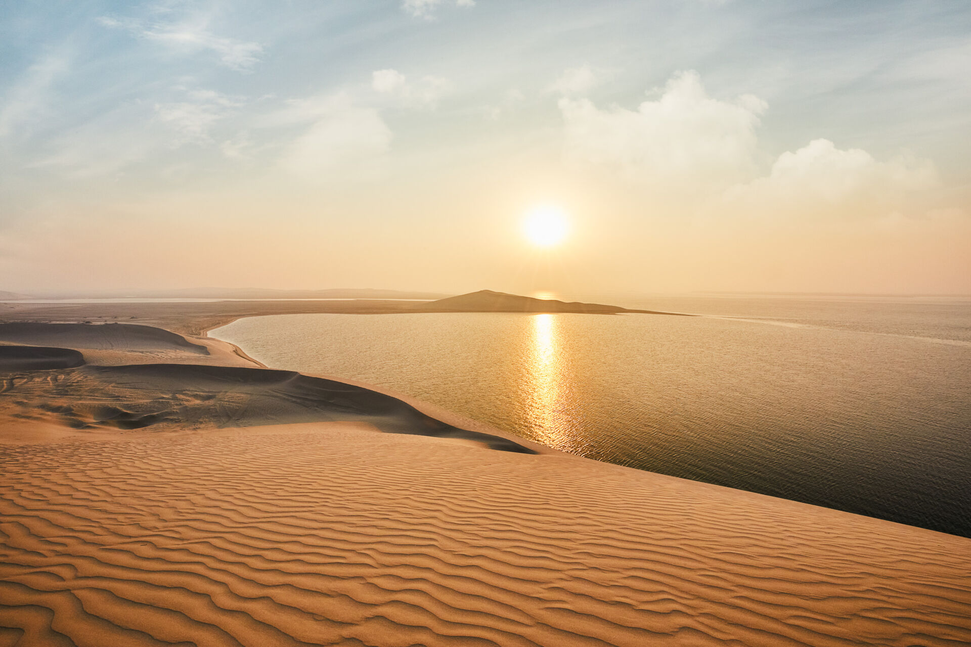 Sand dunes ripple down towards a calm body of water. The dunes curve around a natural bay. It's sunset. This is Khor Al-Adaid, or the Inland Sea.
