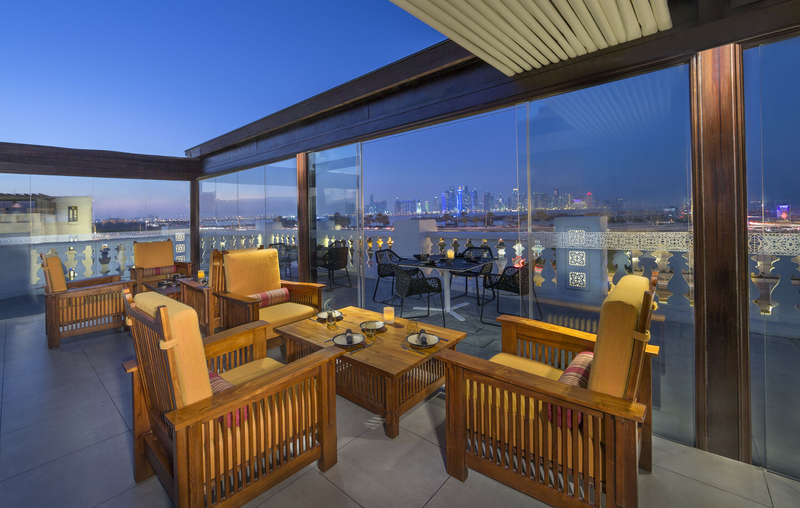 A rooftop restaurant. Inside a glass-walled room with a retractable ceiling are two low wooden tables, each surrounded by three wooden armchairs with cushions on them. Outside the glass wall is a terrace with tables that appear to be metal, and chairs that appear to be some sort of rattan weave. Beyond the terrace, a city skyline is visible, with a cluster of skyscrapers in the centre. It's dusk.