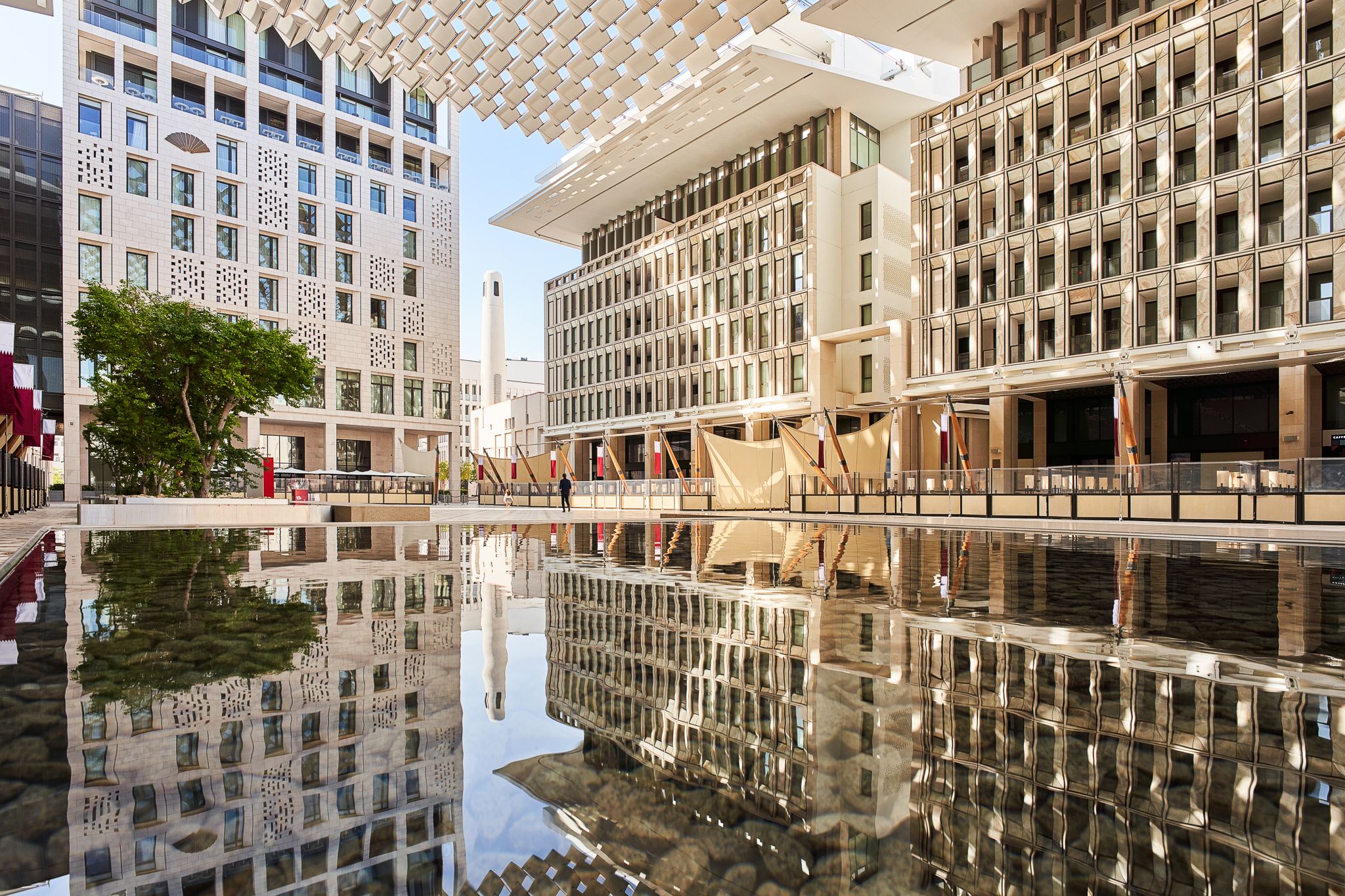 A courtyard. A reflecting pool in the foreground is surrounded by multi-storey buildings with modern architecture. Sun shades are suspended above the courtyard. A minaret is visible in a gap in the buildings in the far corner of the courtyard. Qatari flags hang from the fronts of the buildings. To the left of the reflective pool are a number of leafy trees in a planter. This is Barahat Msheireb.