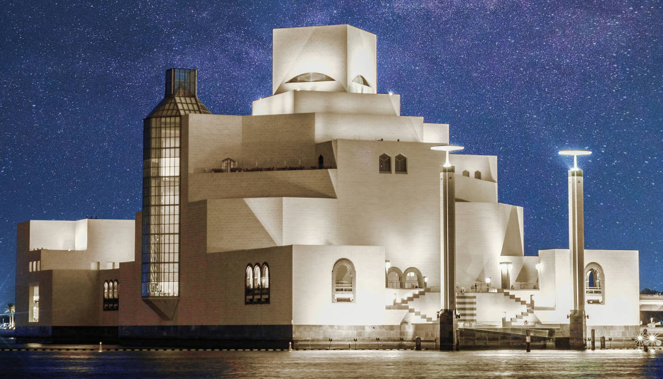 A multi-level building in a modern Middle Eastern architecture style that features blunt walls, sharp angles, returning stairs, few windows and a multi-storey glass tower. Two stone towers topped with lights flank the entrance stairs. It is night time, and a sky full of stars is visible behind the building. This is the Museum of Islamic Art, Doha.
