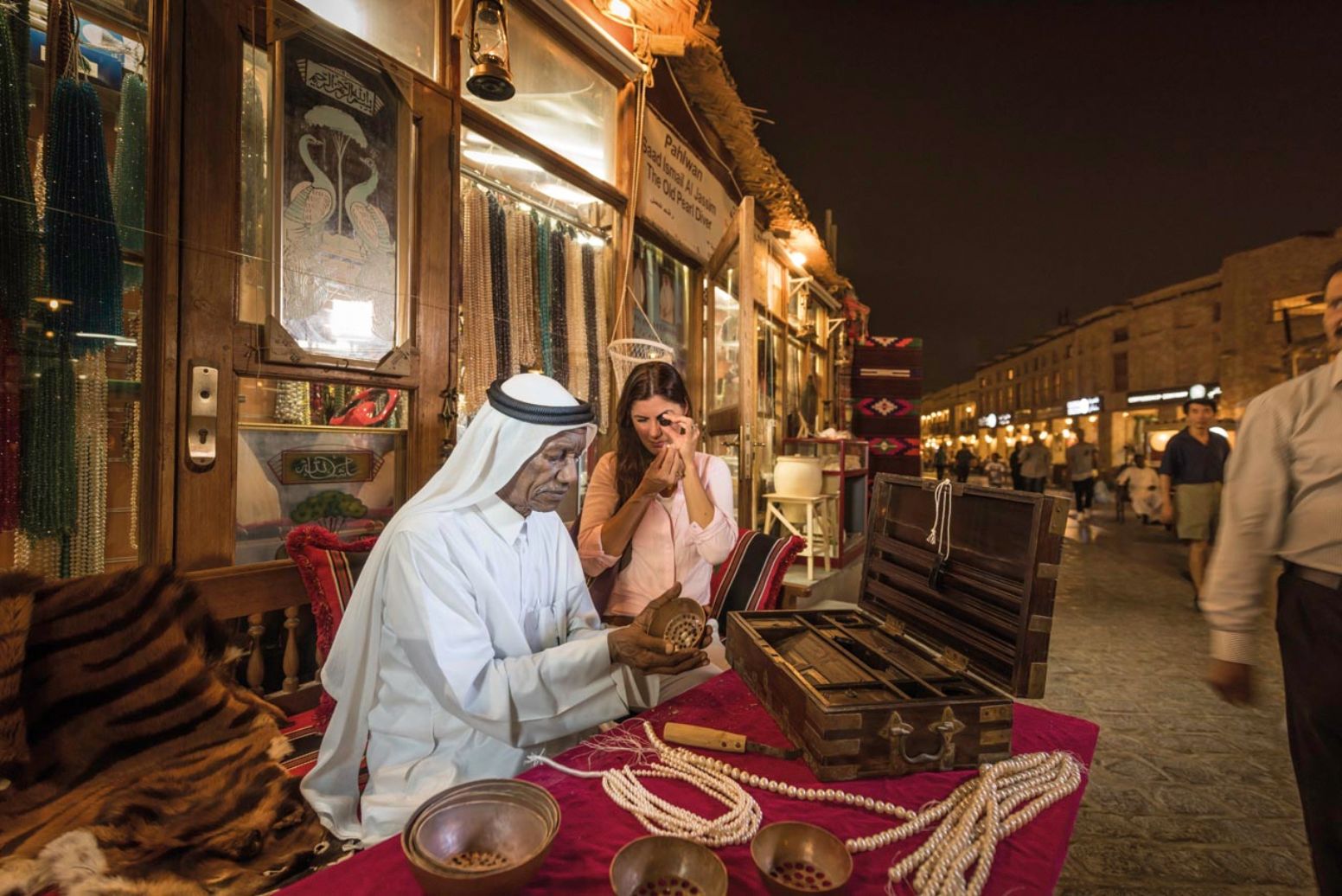 A person sits behind a market stall in Souq Watif selling pearls. Strings of pearls lie on the table in front of them, and they're pouring pearls out of a metal bowl into their hand. A leather case is open on the table. A second person is sitting next to them. This second person is holding a pearl in one hand, and is holding a loupe up to their eye with the other. Behind the pair, more strings of pearls hang from railings. Other stalls stretch away from them into the distance.