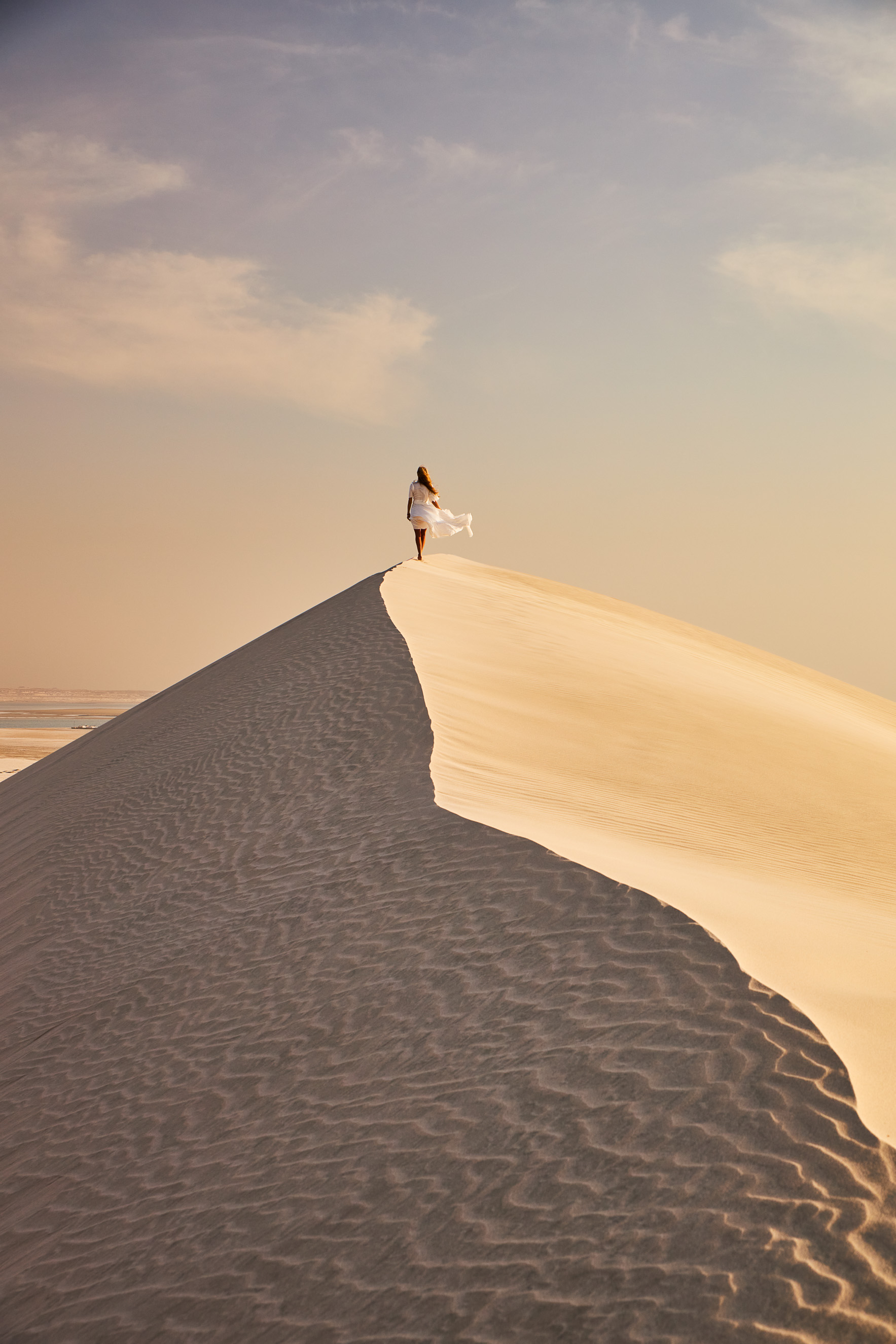 A person with long hair has their back to the camera as they walk along the crest of a sand dune. To their left, the dune is in shadow. Their dress blows in the wind. It is day time. A few thin clouds dot the sky.