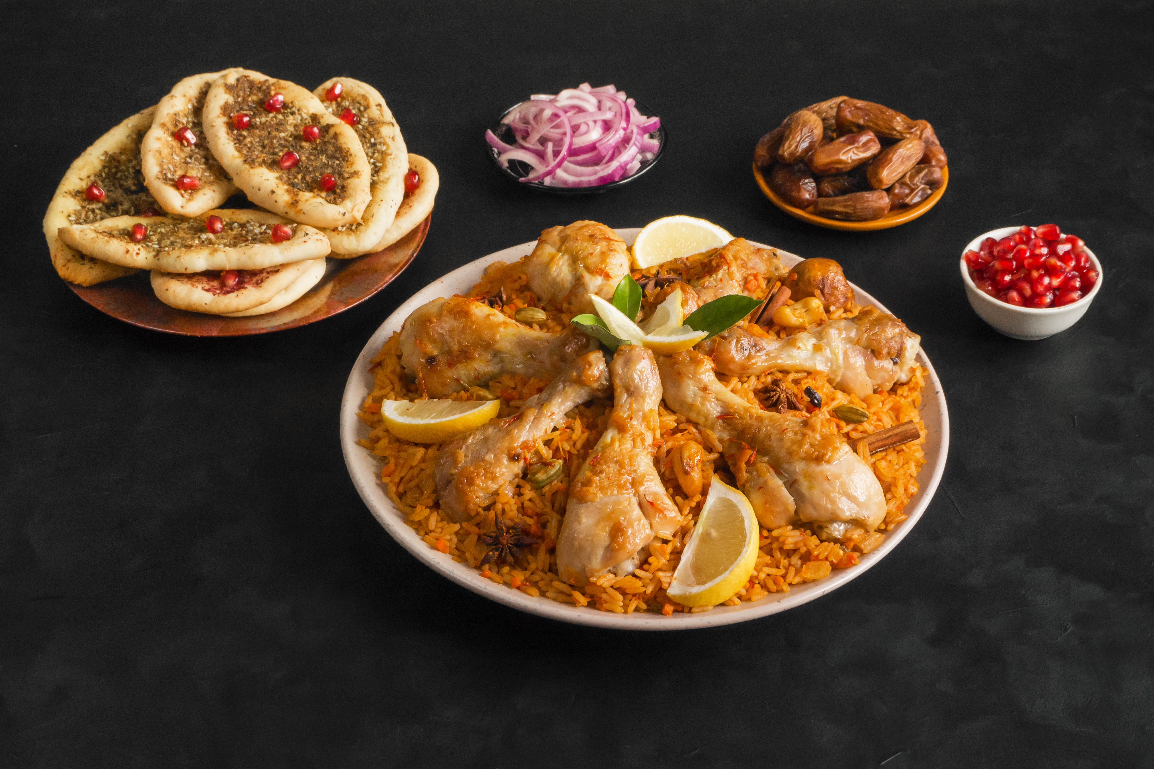 A large plate piled high with spiced rice, chicken legs and lemon wedges (Qatari chicken majboos) in the centre of the frame. Arrayed around the large plate are a smaller plate of flatbreads that are topped with herbs and pomegranate seeds, a dish of chopped onion, a dish of dates, and a dish of pomegranate seeds.