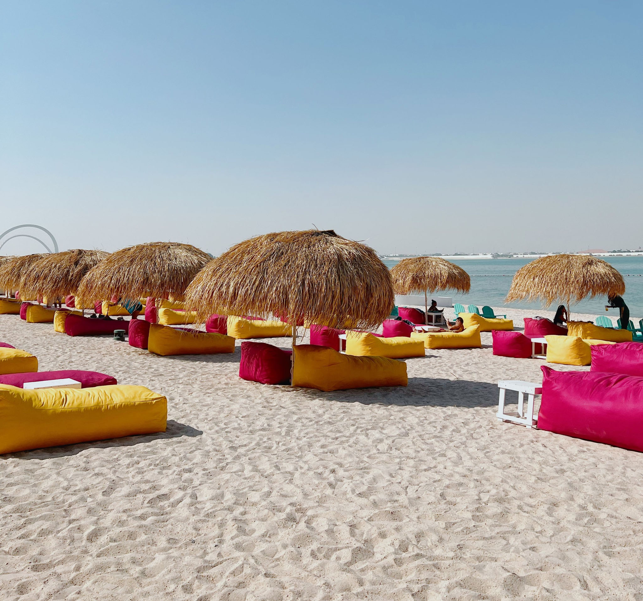 On a sandy beach next to the sea, sun loungers are placed under thatched beach umbrellas. It's day time and the sky is clear except for one thin cloud.