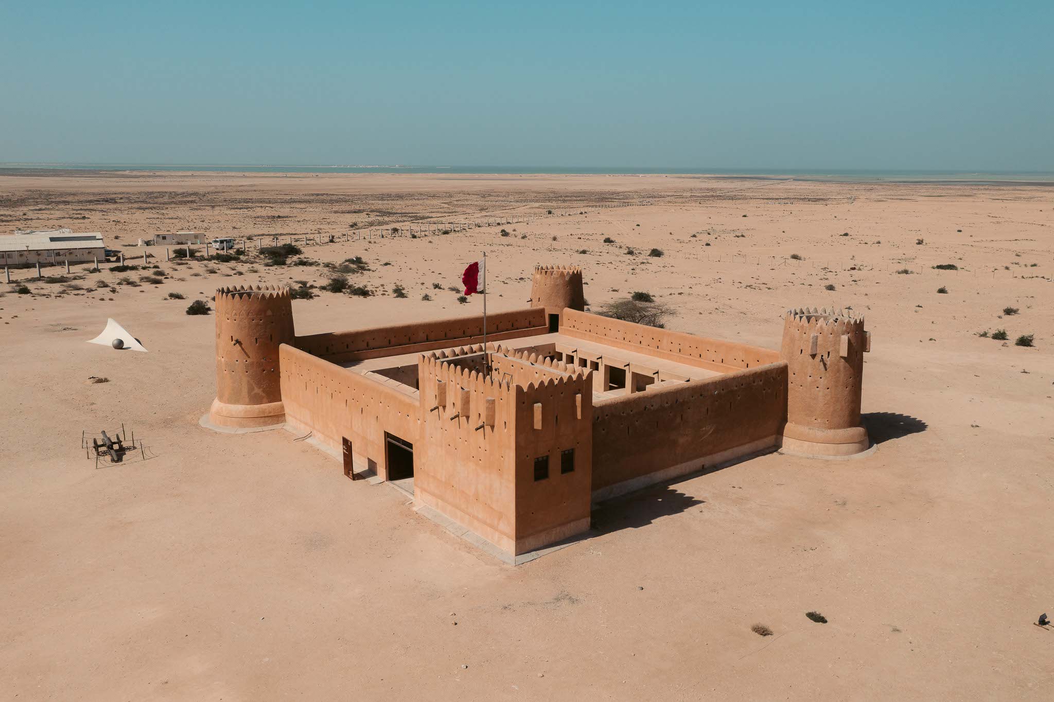 A square fortress (Al Zubarah) surrounded by bare sandy earth. Round towers stand at three of the fortress's corners. The fourth corner, next to the fortress's entrance, is dominated by a rectangular tower, from which the Qatari flag flies. Outside the walls of the fortress sits a small cannon, surrounded by ropes. A wire fence stretches behind the fortress. On the other side of the fence is a low modern building and a bus. A sliver of sea is visible on the horizon. It's day time.