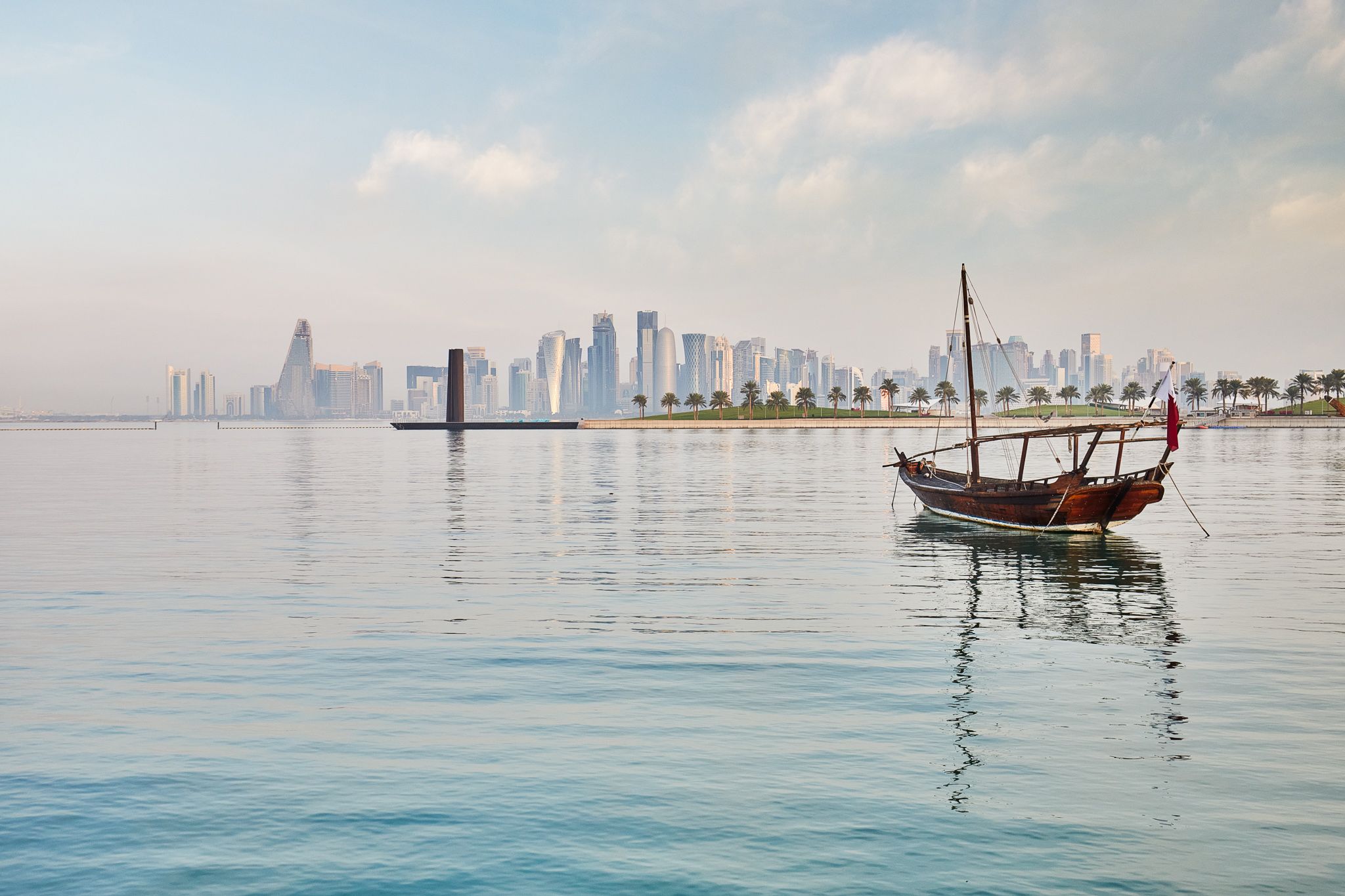 An empty wooden boat (a dhow boat) anchored in a calm bay. The boat has a mast and a shaded area open on the sides, and a Qatari flag flies from the stern. In the middle distance, a row of palm trees lines a waterfront, leading to the famed 7 sculpture. Behind 7, the skyscrapers of Doha are visible. It's day time.