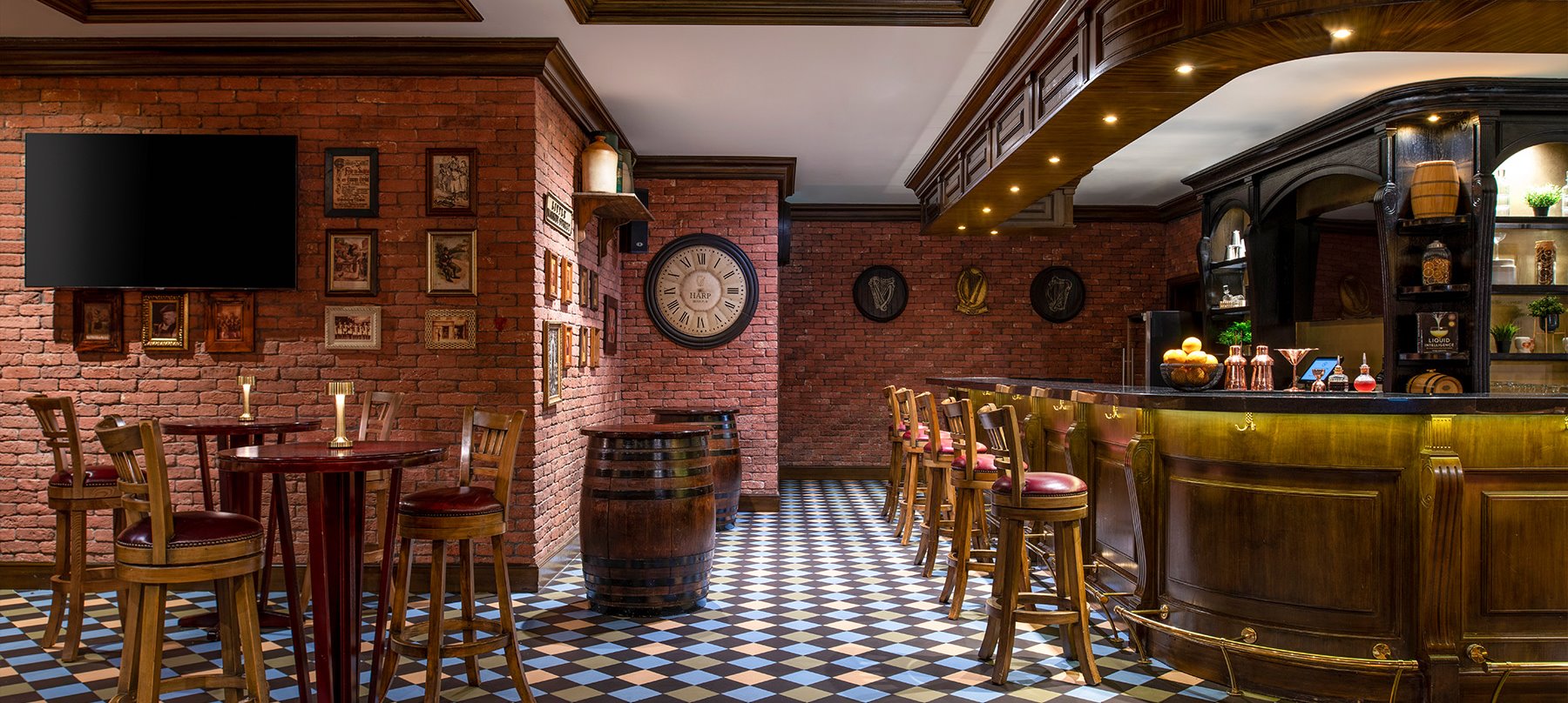 Bar interior. A checkered tile floor. Red brick walls, covered with pictures in old-style frames and illustrations of the Irish harp. A TV hangs on the wall in the left of the image. Two wooden tables with high chairs on the left, and two barrels that have been turned into tables in the centre. To the right is the bar itself, which is wooden, and has a brass foot rail and brass hooks for coats and bags. Five high chairs are lined up in front of the bar. A bowl of lemons sits on the bar next to cocktail-making equipment (shakers, a strainer and a jigger).