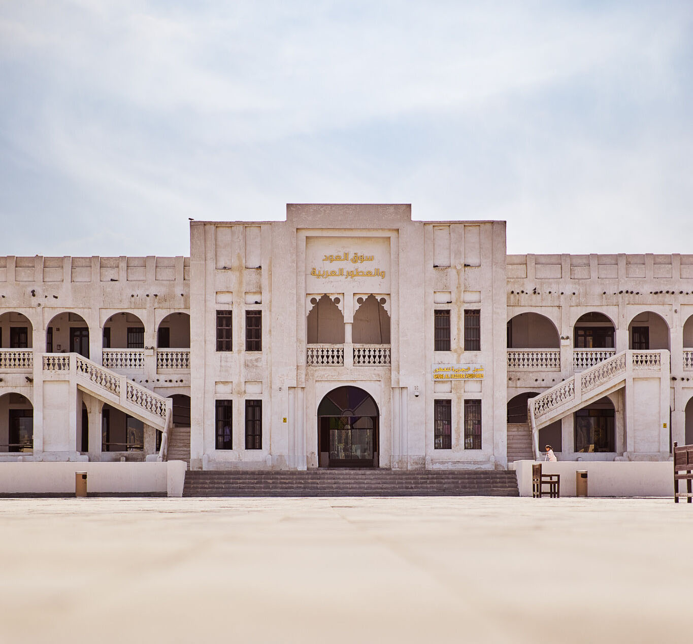 A squat two-storey building viewed from across a tiled courtyard. The architectural style is modern Middle Eastern, with covered colonnades on both floors and a flat roof. A short, broad flight of stairs leads up to glass front doors. It's day time.