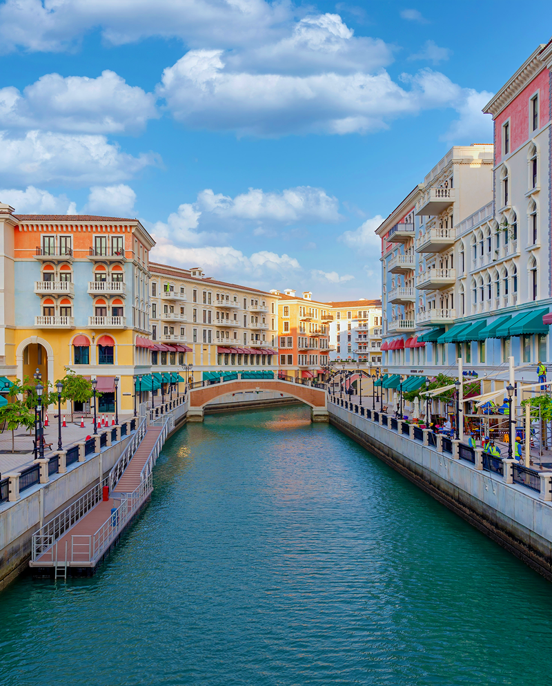 A canal flows between Italian-style multi-storey buildings with balconies and window awnings. A humpback bridge spans the canal. On both banks are spacious walkways. It is day time. This is Qanat Quartier.