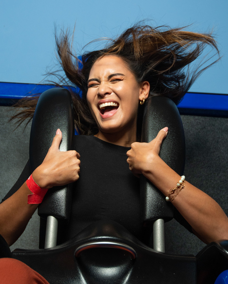 A person sits on an amusement ride. They are strapped in with an over-the-shoulder harness, which they're holding with both hands. Their eyes are closed and they're laughing as their hair whips around in the wind.