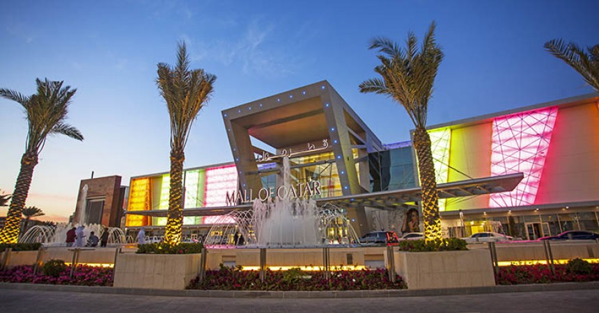 Exterior of a large modern building. A sign above the entrance reads 'Mall of Qatar'. In front of the building are several fountains and a row of palm trees. It's dusk.