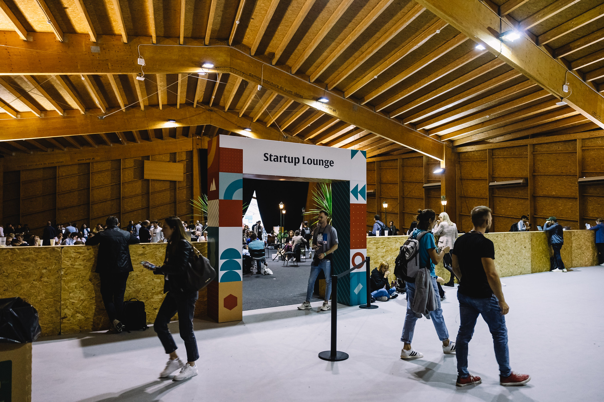 People walk in front of a low chipboard wall. A person stands under an arch in the centre of this wall. This is an entryway. Text on the arch reads 'Startup Lounge'. Beyond the wall and arch, groups of people are seated at small tables. The space looks industrial, with exposed chipboard walls, and a chipboard ceiling with wooden support beams.