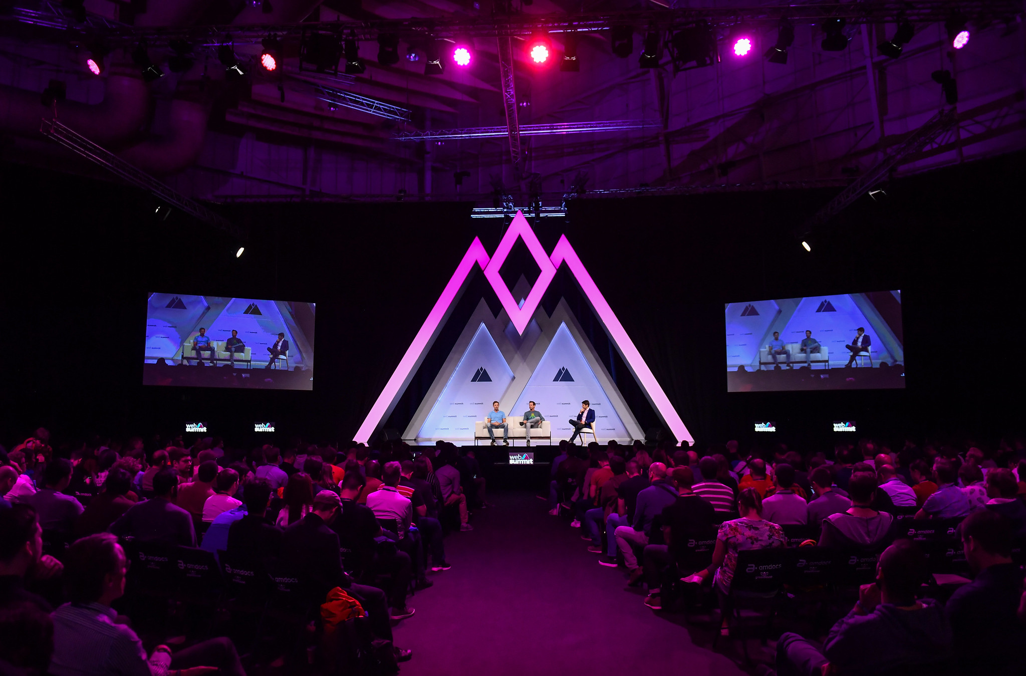 Photo of a lit up stage at Web Summit. The stage has mountain-shaped lights. There appear to be three speakers sitting on the stage engaged in conversation. There is a full audience watching the stage. There are two screens on either side of the stage.