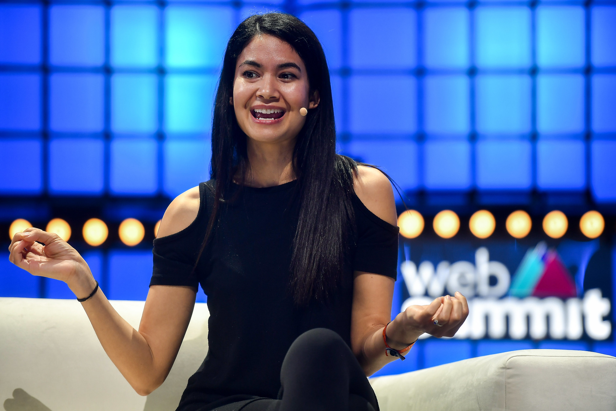 Photograph of a person (Melanie Perkins, co-founder and CEO of Canva) on stage at Web Summit. The person is sitting on a chair with their hands lifted on either side of them. The Web Summit logo is visible on the stage background behind the person.