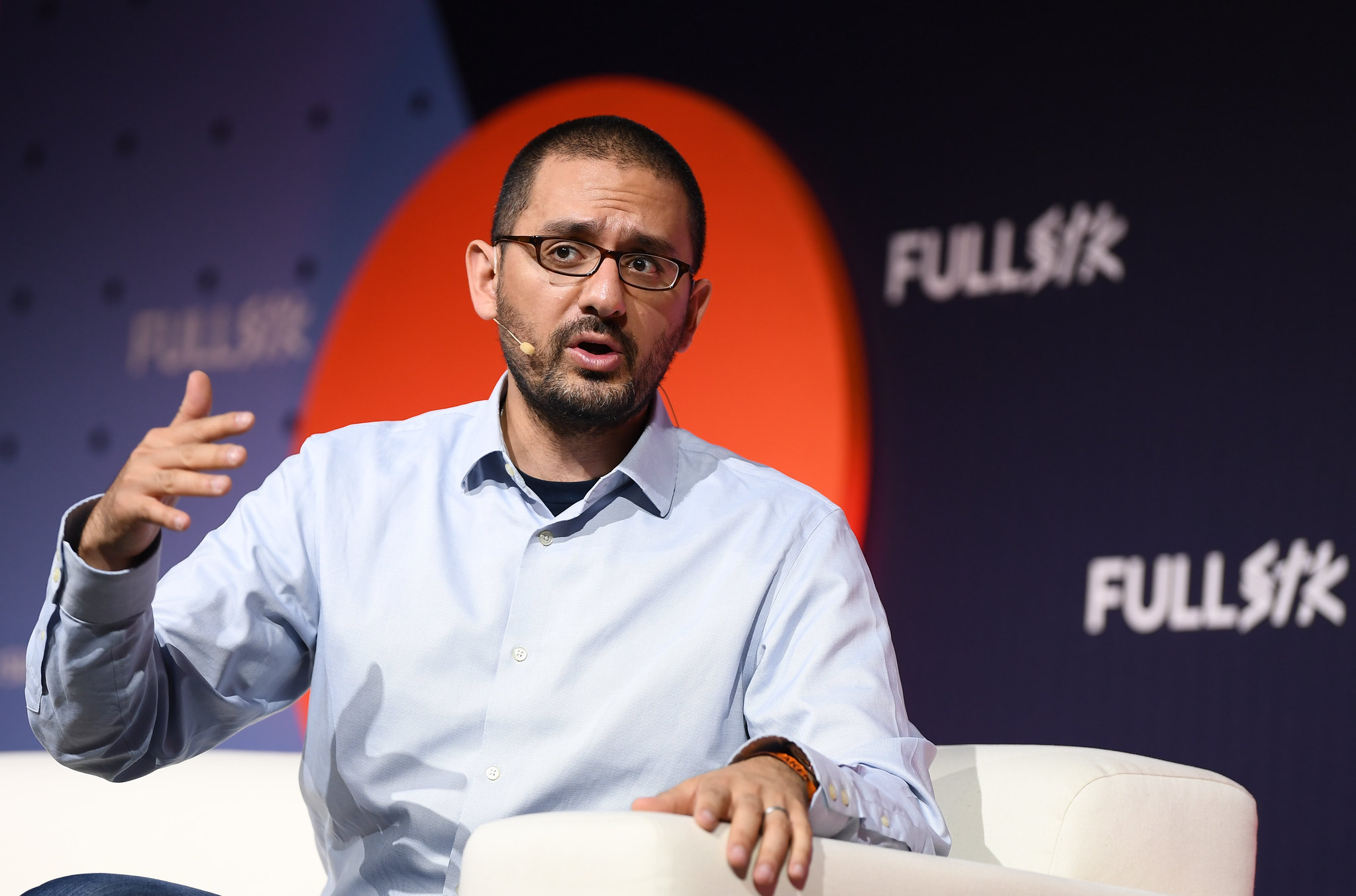 Photo of a person (Raffi Krikorian, former CTO of the Democratic National Committee (DNC)), speaking on stage at FullSTK. They are sitting on a chair and gesturing with their hands. They are wearing an over-ear microphone.