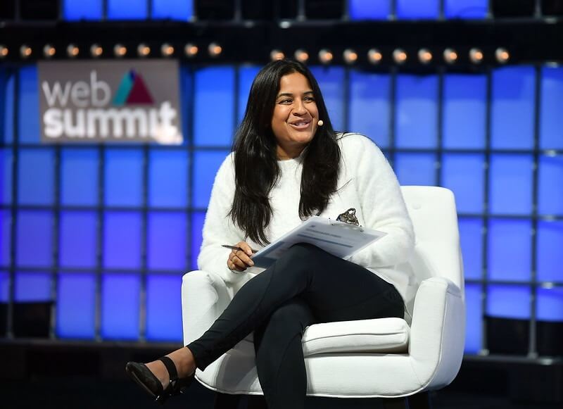 A person (Seedcamp managing partner Reshma Sohoni) sits in an armchair. The person's left leg is crossed over their right, and a clipboard is balanced on their knee. They're holding a pen in their right hand, and wearing a radio mic. They appear to be speaking. The Web Summit logo is visible over their right shoulder.
