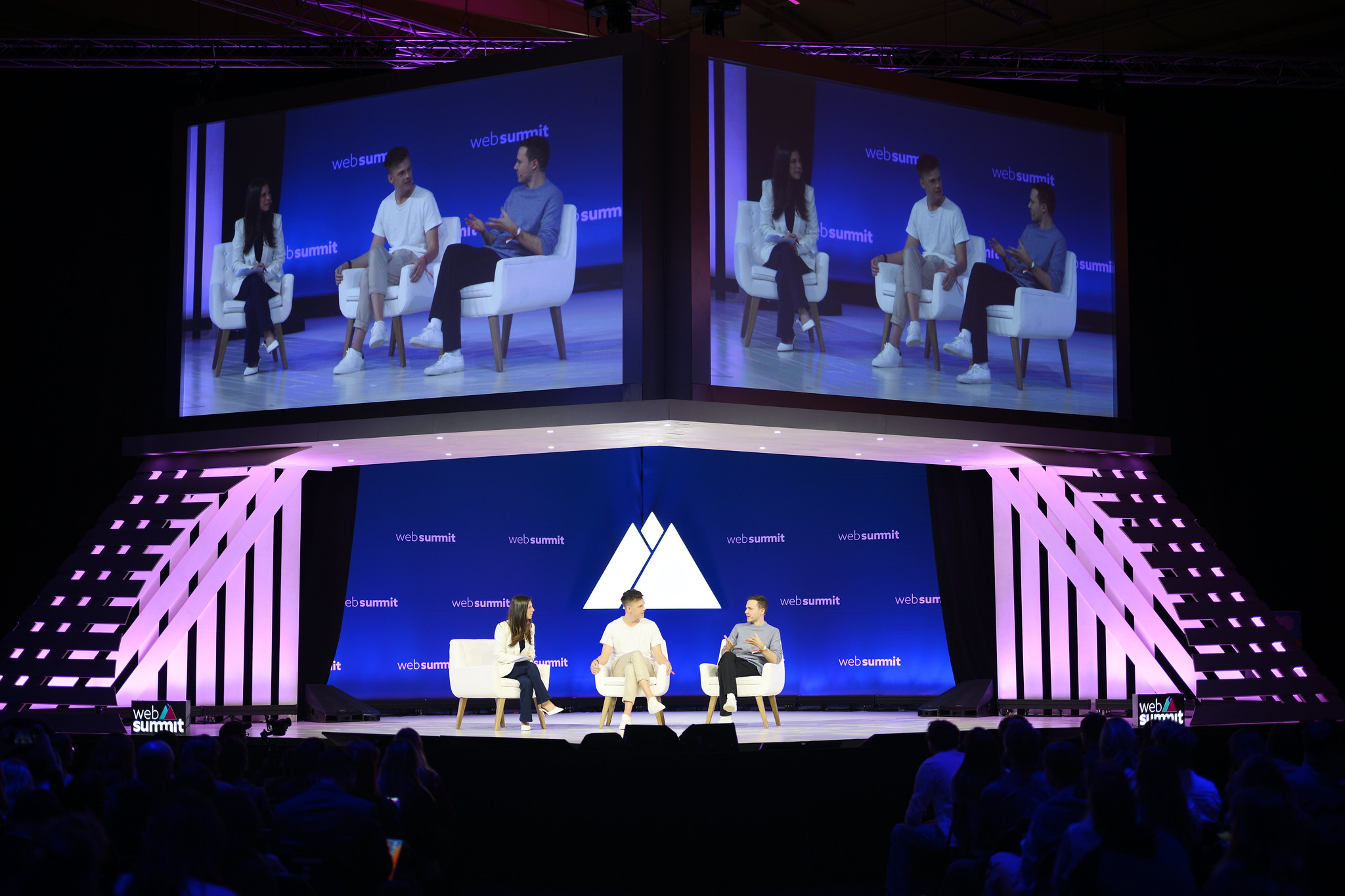 An event stage viewed over the heads of a packed audience. Three people sit on stage. Two large screens over the stage show a close-up of these people. The Web Summit logo is visible behind the people.