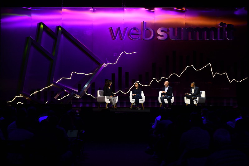 Zoomed out photo of four speakers on stage at Web Summit. The four speakers are sitting on chairs. There appears to be an audience of people watching them. In the background, the Web Summit logo is visible. The stage is dark and there are neon lights above the speakers.