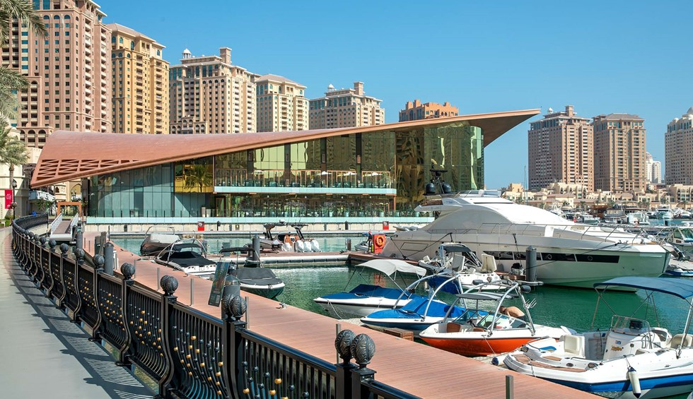 A marina where several boats and yachts of varying sizes are docked. A modern building alongside the marina sports floor-to-ceilings windows over two storeys. Behind this building are several high-rise buildings.