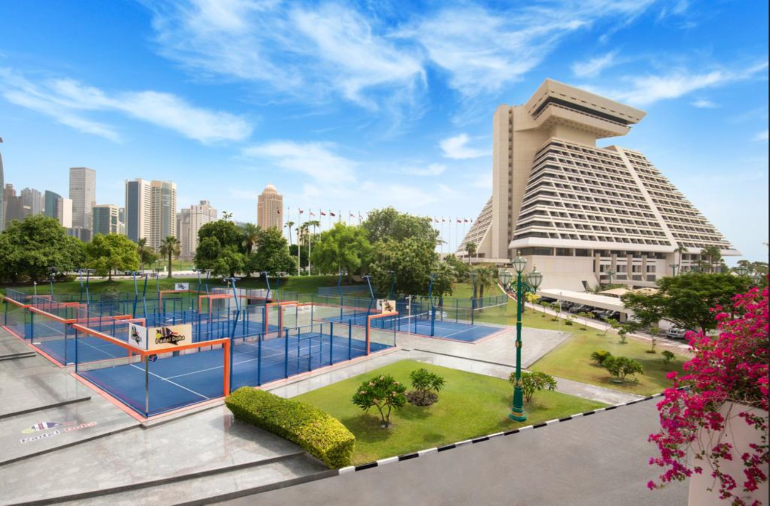 An artist's rendering of several padel courts with fenced perimeters. They are next to a modern multi-storey pyramid-shaped building. In the background is a dense cluster of modern high-rise buildings. The courts and building are surrounded by grass and trees. Thin clouds dot the sky.