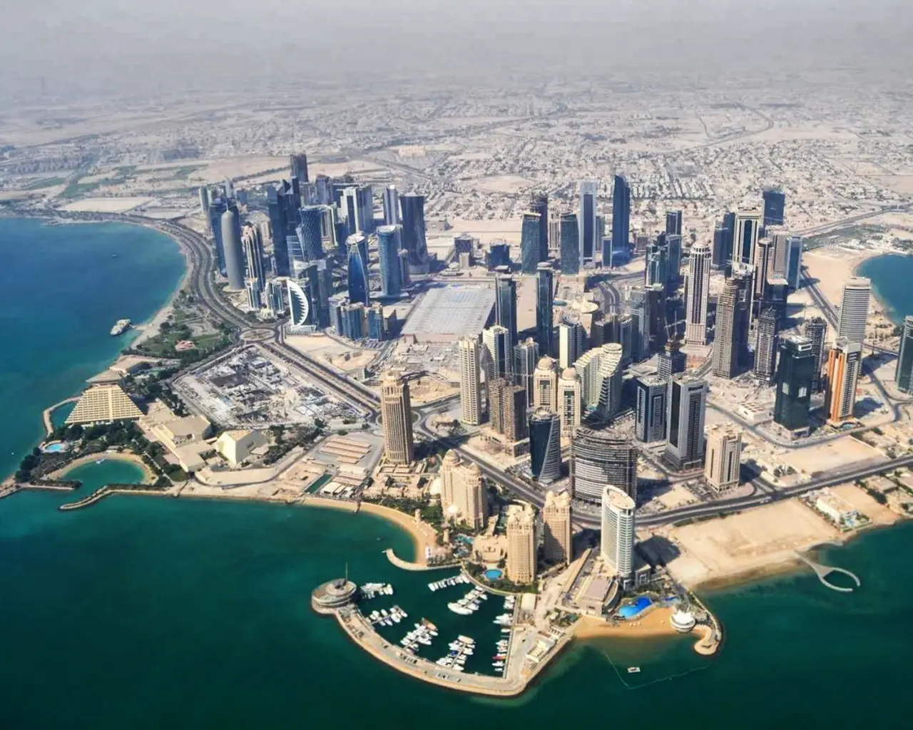 An aerial view of Doha's West Bay area. Dozens of high-rise buildings are clustered on a short peninsula. Where the lands meets the water, there are beach areas and marinas.