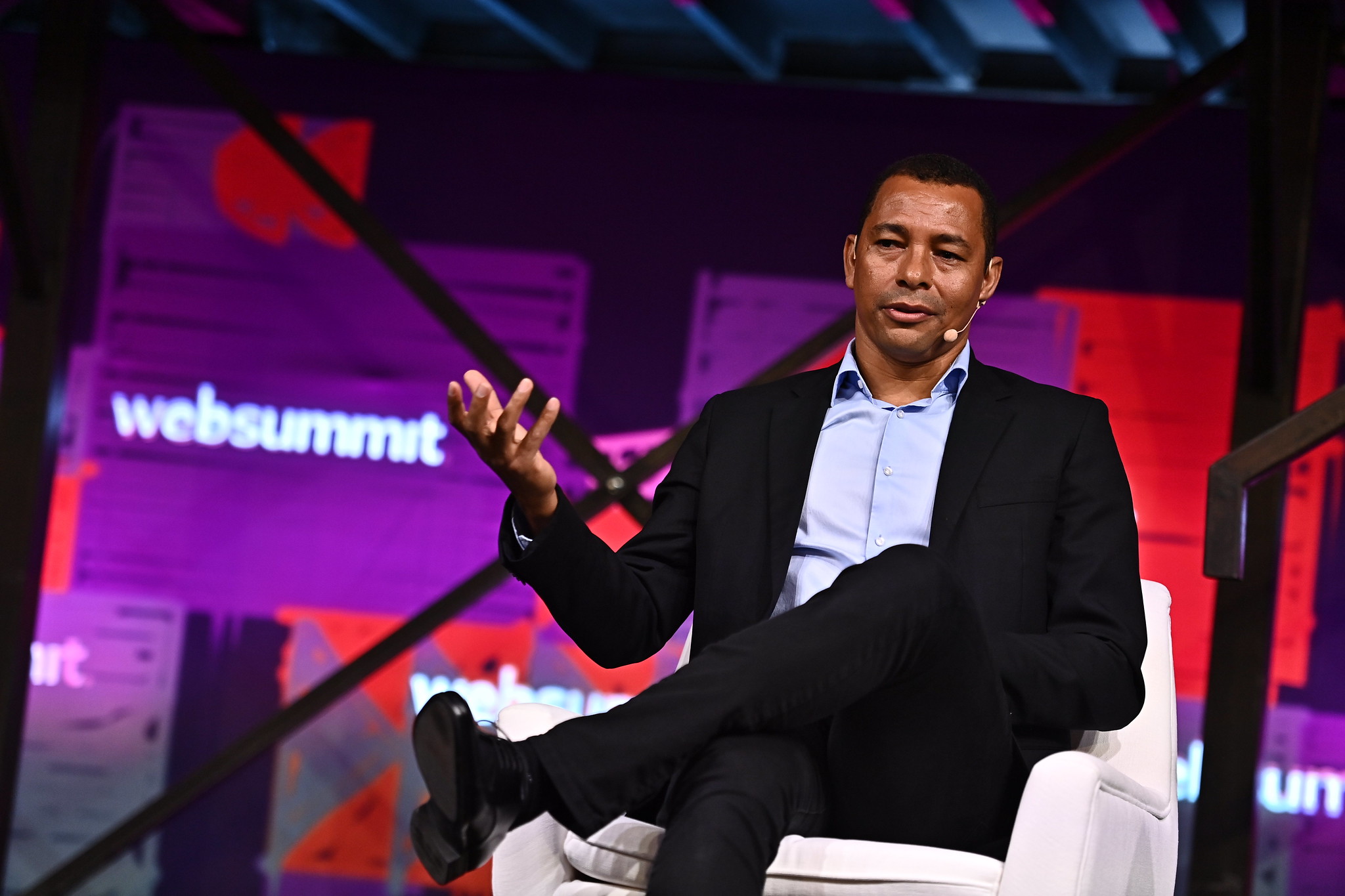 Photograph of World Cup Winner Gilberto Silva on stage at Web Summit. Gilberto is sitting on a chair and has one hand lifted as if asking a question. The Web Summit logo is visible in the background.