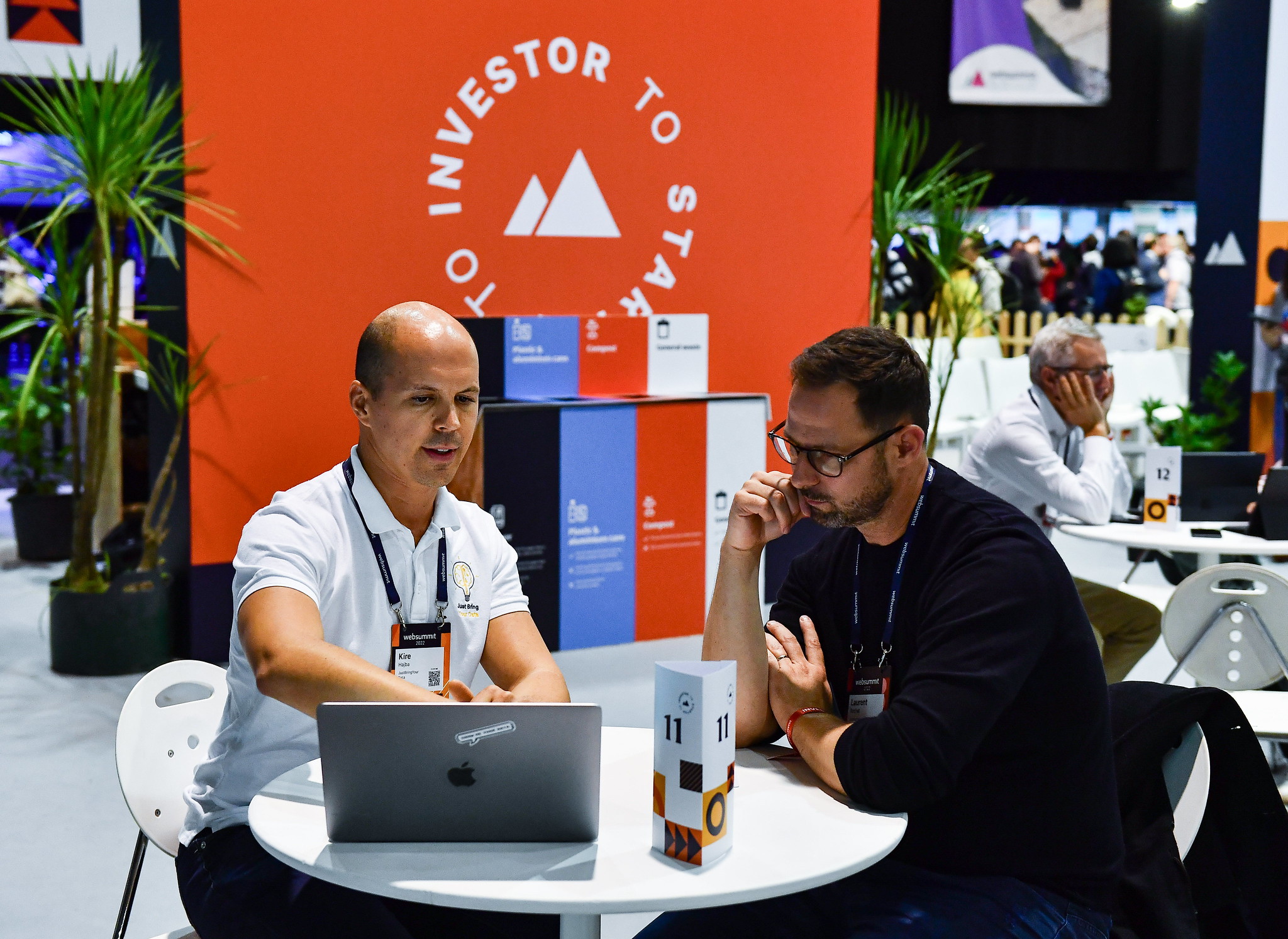 Two people sit at a round table, looking at an open laptop. One of the people is pointing to the laptop screen with their right hand, and letting their left forearm rest on the table. The other is resting their right forearm on the table, and has their right hand raised towards their mouth in a manner that suggests focus. Behind them, investor to startup is written in a circle around the Web Summit logo. Below the logo is a bank of rubbish bins. To the left of the logo is a small potted palm tree.