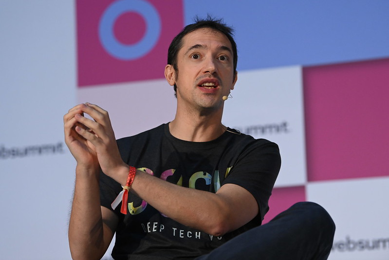 Oscar Ramos speaks on the Venture Stage at Web Summit. Oscar is sitting down and speaking, clasping their hands together and wearing a microphone. The Web Summit logo appears on several parts of the stage background.
