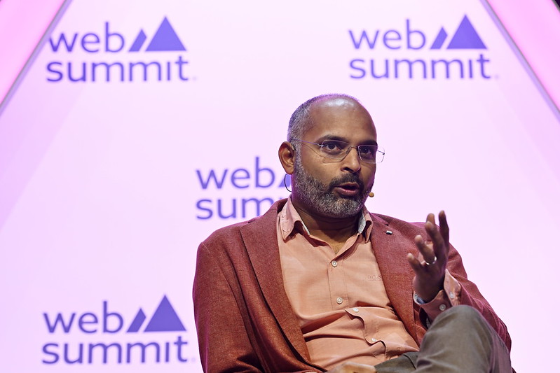 A person (Zopa CEO Jaidev Janardana) sits in an armchair. They are leaning on the right armrest of the armchair. They are wearing a headset mic and gesturing with their left hand. They appear to be speaking. The Web Summit logo is visible in several places behind them. This is onstage at Web Summit 2023.