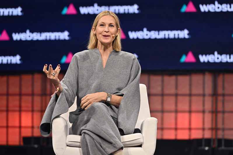 A person (actor and Whyzzer investor Kelly Rutherford) sits in an armchair. Their left arm is crossed over their lap, and they're gesturing with their right hand. They're wearing a headset mic and are smiling. They appear to be speaking. Behind them, the Web Summit logo is visible in several places. This is Web Summit 2023, Centre Stage.