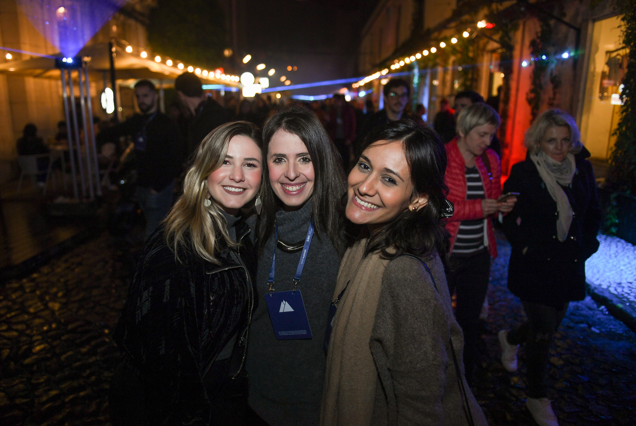 Three smiling people hug each other and look directly into the camera lens. The person in the centre is wearing a lanyard with the Web Summit logo. Behind them is a cobbled street full of people. It's night time. This is Night Summit at LX Factory in Lisbon.