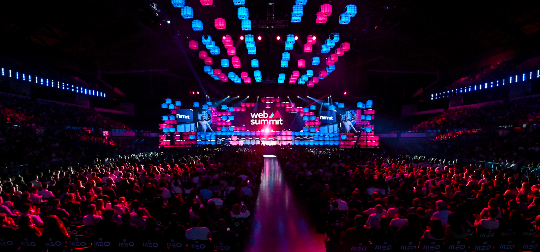 A large stage in an arena, viewed over the heads of a packed audience. A broad aisle runs up the middle of the crowd towards the stage. The Web Summit logo is visible on the stage.