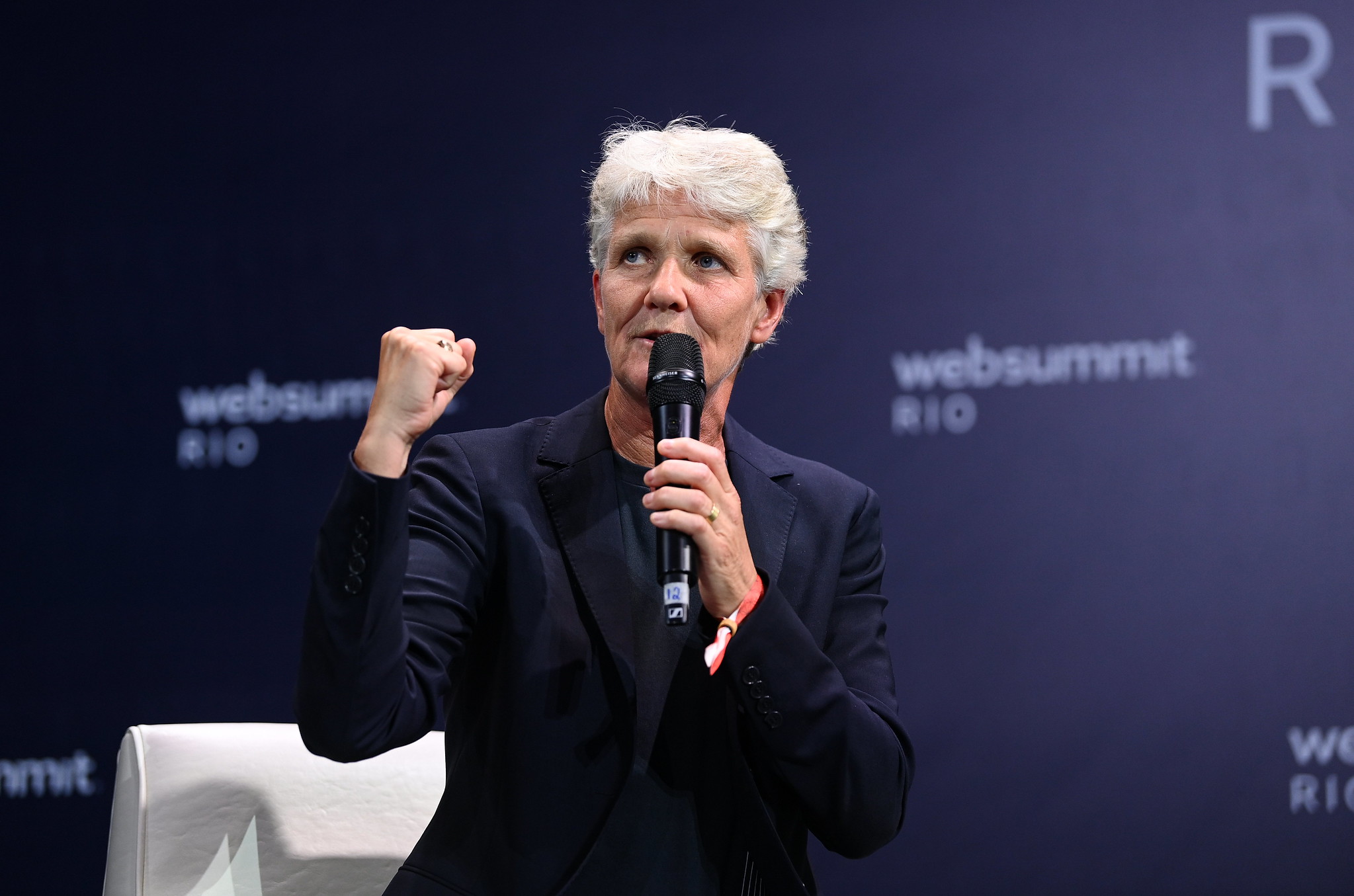 Photograph of a person (Pia Sundhage, former head coach of Brazil women's national team) speaking on stage at Web Summit Rio. The person is holding a microphone and has their fist in the air. They are sitting on a chair.