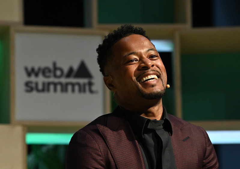 A person (former footballer Patrice Evra) pictured from the chest up. They are wearing a headset mic and laughing. Behind them is a backdrop made of wooden boxes. The Web Summit logo is visible over their right shoulder. This is Web Summit's Q&A stage.