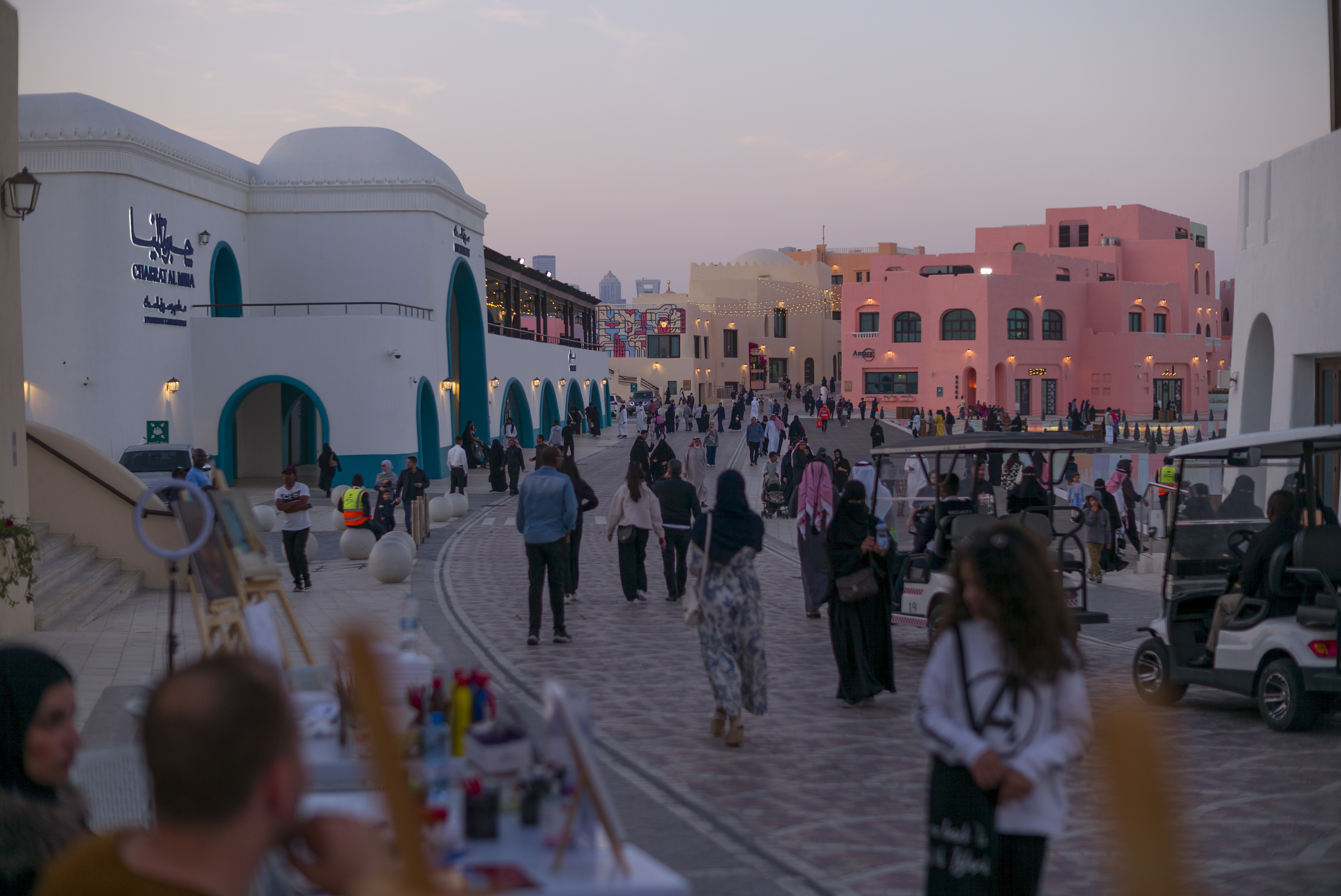 A bustling marketplace, surrounded by stone buildings. People walking about the street, some are standing to observe paintings at an outdoors stall. This is Souq Waqif.