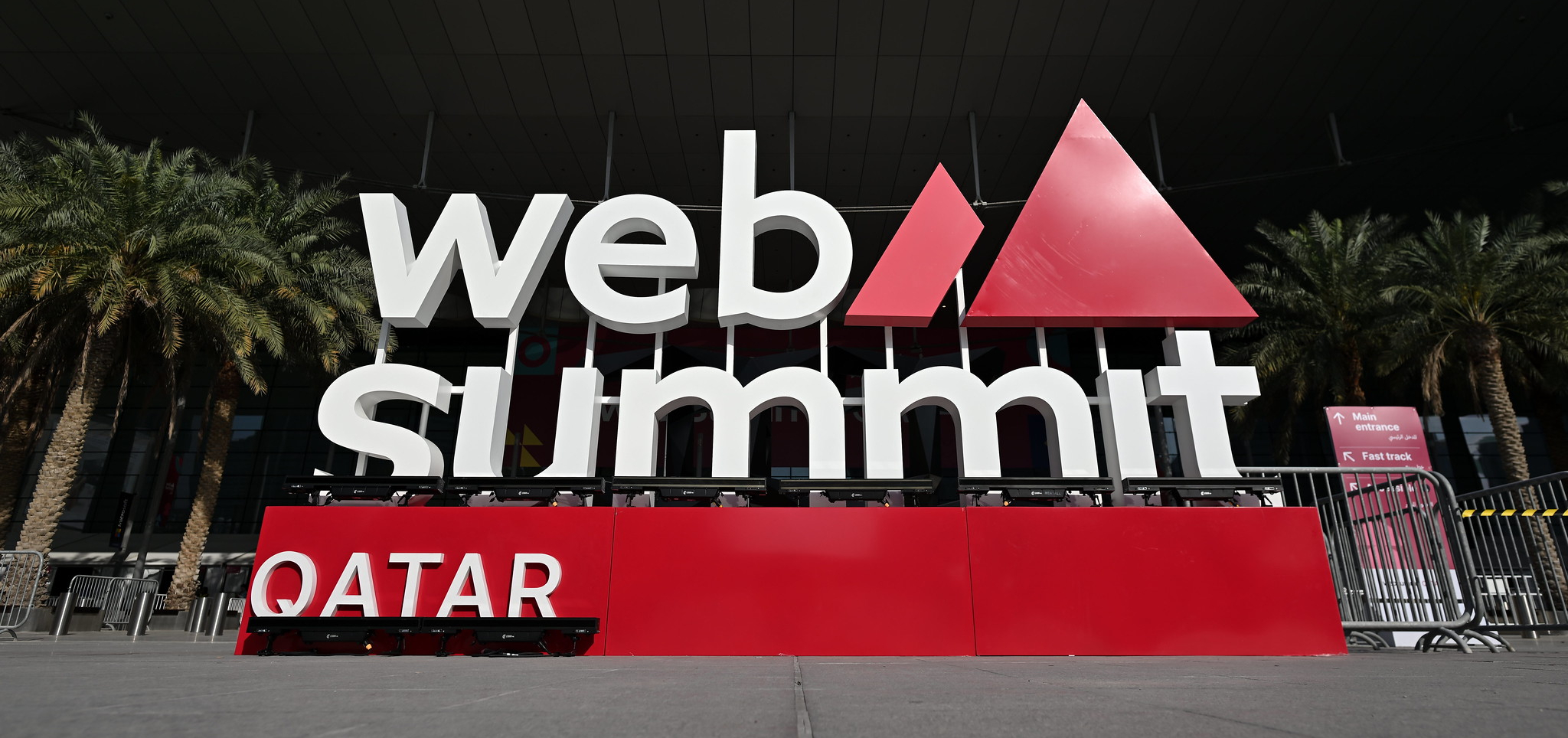 A large sign in the shape of the Web Summit Qatar logo. Behind the sign are a number of palm trees, standing in front of a modern conference centre. There are no clouds in the sky.