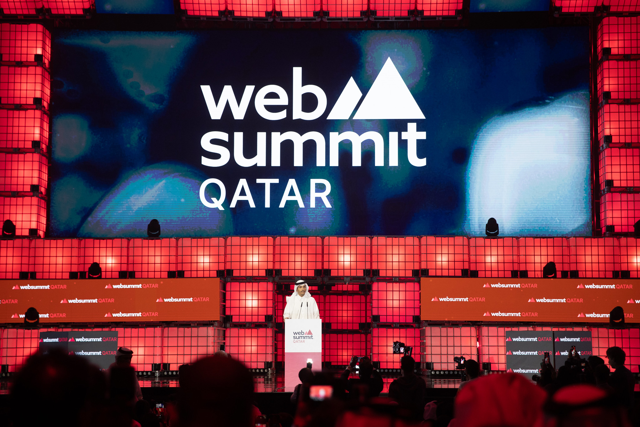 A large stage viewed over the heads of a packed audience. The Web Summit Qatar logo is visible on a large screen that dominates the back wall of the stage. In the centre of the stage stands His Excellency Sheikh Mohammed bin Abdulrahman Al Thani, the State of Qatar's prime minister and minister of foreign affairs