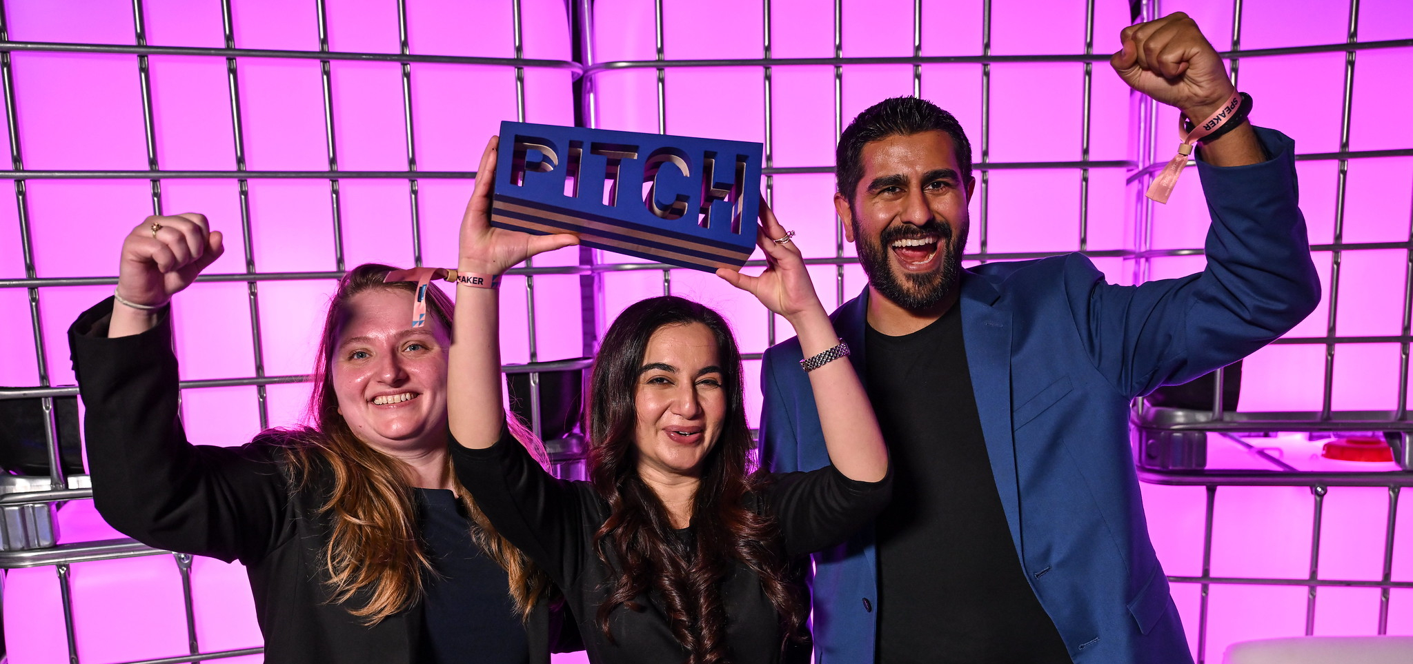 Three smiling people facing the camera raise their arms above their heads in apparent celebration. The person in the middle is holding a wooden trapezoidal trophy with the word PITCH laser cut in it. These are the winners of PITCH at Web Summit Qatar. In the centre is Breshna founder and CEO Mariam Nusrat. On the left is Breshna VP Joanna Kurylo. On the right is Blockchain Founders Fund managing partner Aly Madhavji, an investor in Breshna.