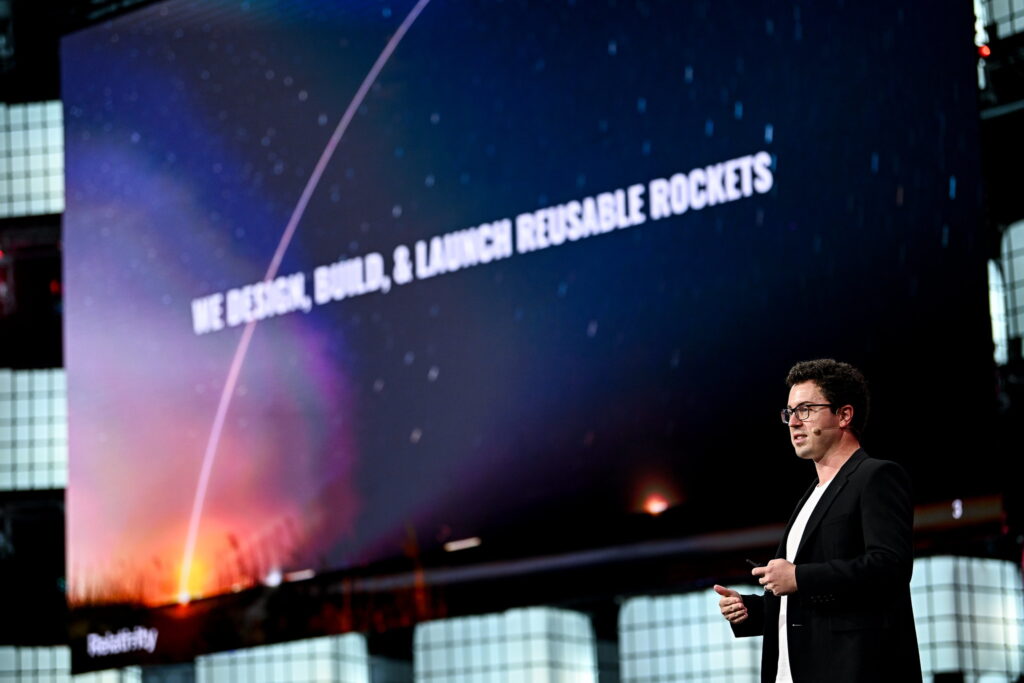 A person (Relativity Space co-founder and CEO Tim Ellis) stands on stage. Tim is wearing a headset mic and appears to be speaking. Behind Tim, a large screen bears the message 'we design, build and launch reusable rockets'. This is Centre Stage at Web Summit Qatar.