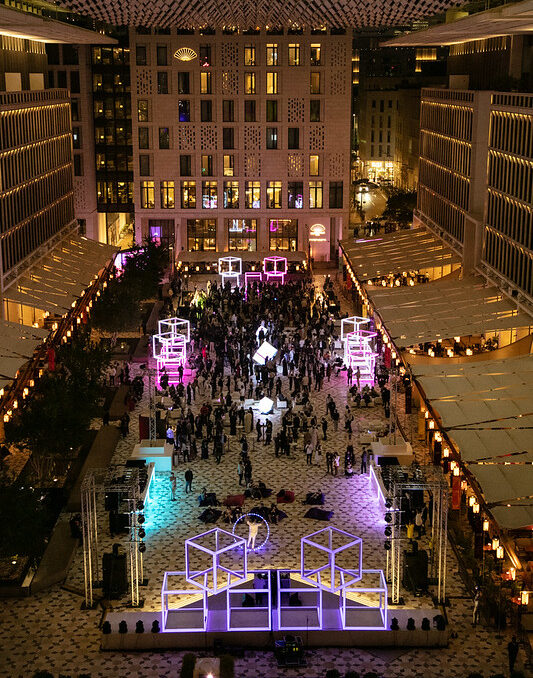 Birds-eye photograph of Night Summit at Barahat Msheireb. There is a crowd of people in the venue. There are lights running along the venue, and there are lit-up squares at the top.
